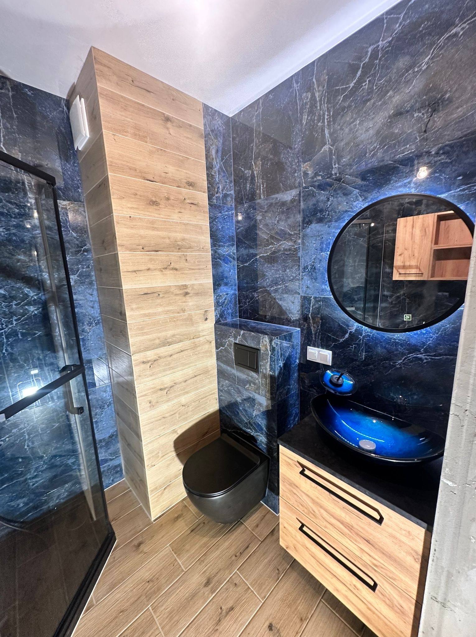 Niebiesko-marmurkowa łazienka z drewnianymi akcentami: szafka pod umywalkę, zabudowa WC, podłoga. Okrągłe lustro z podświetleniem LED i szklana kabina.