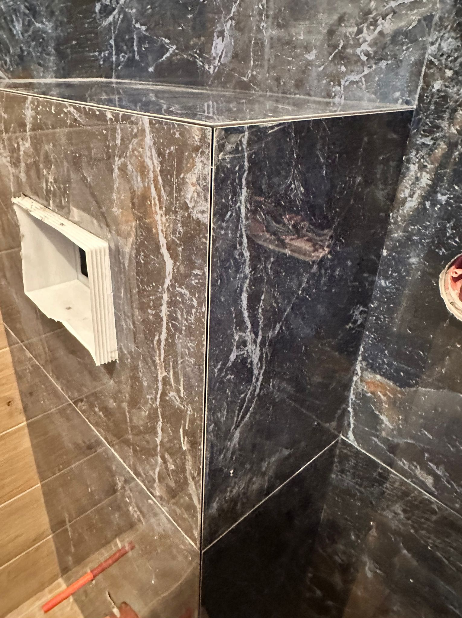 Wykończona obudowa z płytek imitujących marmur, z widocznym otworem wentylacyjnym i fragmentem podłogi. Detal wykończenia narożnika.