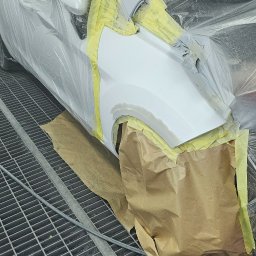 2KB AUTO DETAILING - Biały samochód przygotowany do lakierowania, oklejony taśmą i folią ochronną, widoczna kratka podłogowa lakierni.