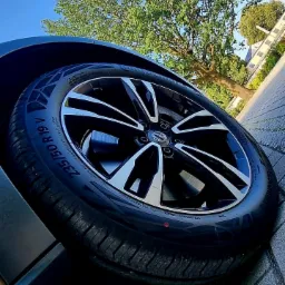 Błyszcząca, czarna felga aluminiowa z oponą 235/50 R19 V na tle błękitnego nieba i korony drzewa.