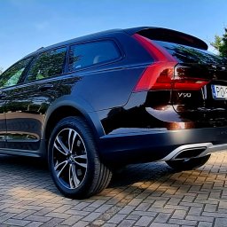 2KB AUTO DETAILING - Ciemnobrązowy samochód Volvo V90 Cross Country, widziany z tyłu pod kątem, zaparkowany na kostce brukowej na tle zieleni i błękitnego nieba.