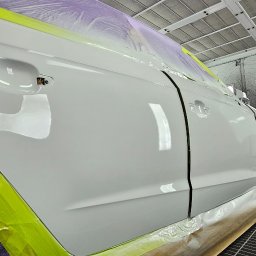 2KB AUTO DETAILING - Biały samochód w kabinie lakierniczej, zabezpieczony folią i taśmą, z widoczną świeżo polakierowaną karoserią i maską czekającą na montaż.