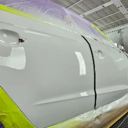 Biały samochód w kabinie lakierniczej, zabezpieczony folią i taśmą, z widoczną świeżo polakierowaną karoserią i maską czekającą na montaż.