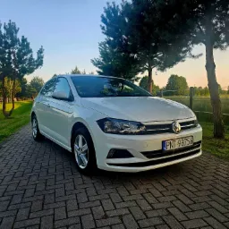 Biały samochód Volkswagen Polo zaparkowany na brukowanej drodze z widokiem na zielone drzewa i pole w tle o zachodzie słońca.