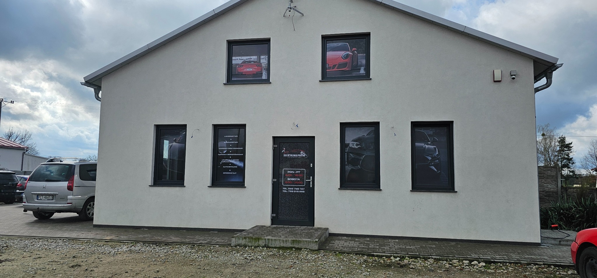 Elewacja budynku z oknami, w których widoczne są plakaty samochodów oraz szyld z napisem '2KB Detailing & Painting', informujący o usługach detailingowych i lakierniczych.