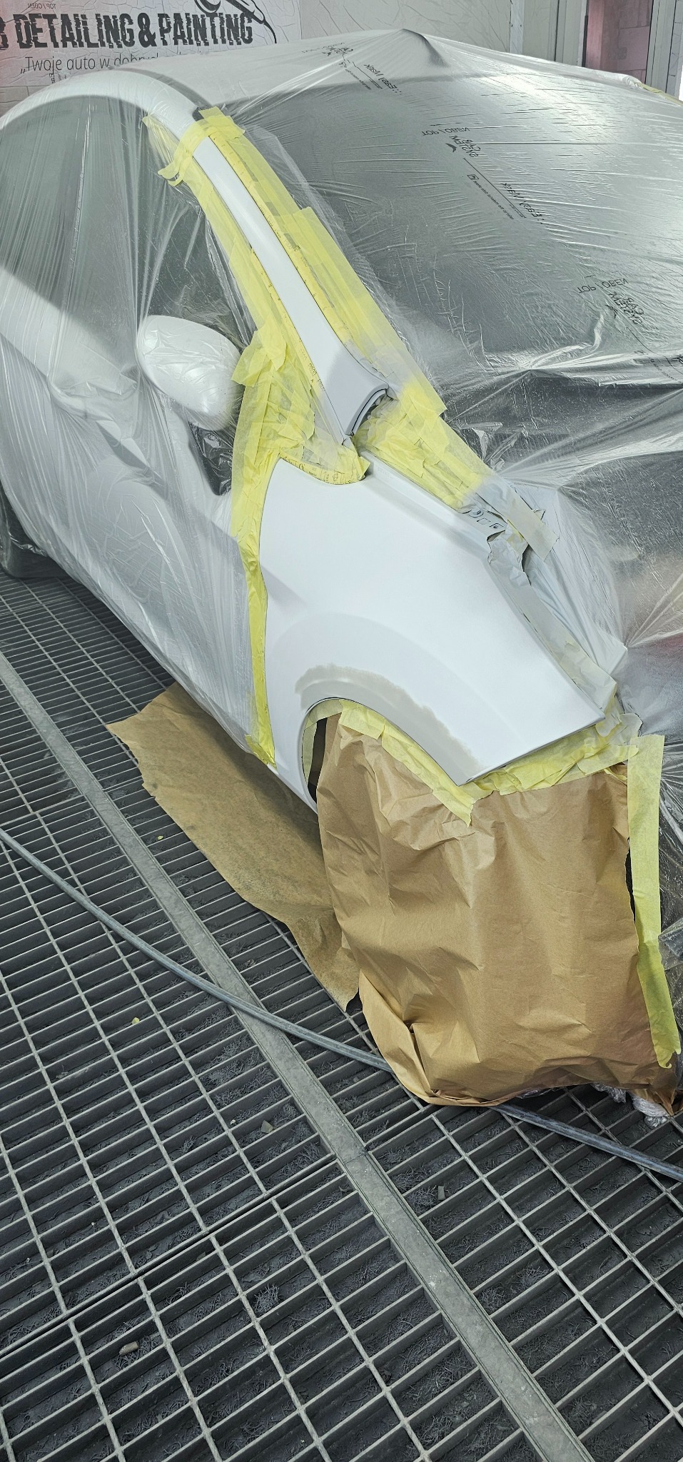 Biały samochód przygotowany do lakierowania, oklejony taśmą i folią ochronną, widoczna kratka podłogowa lakierni.