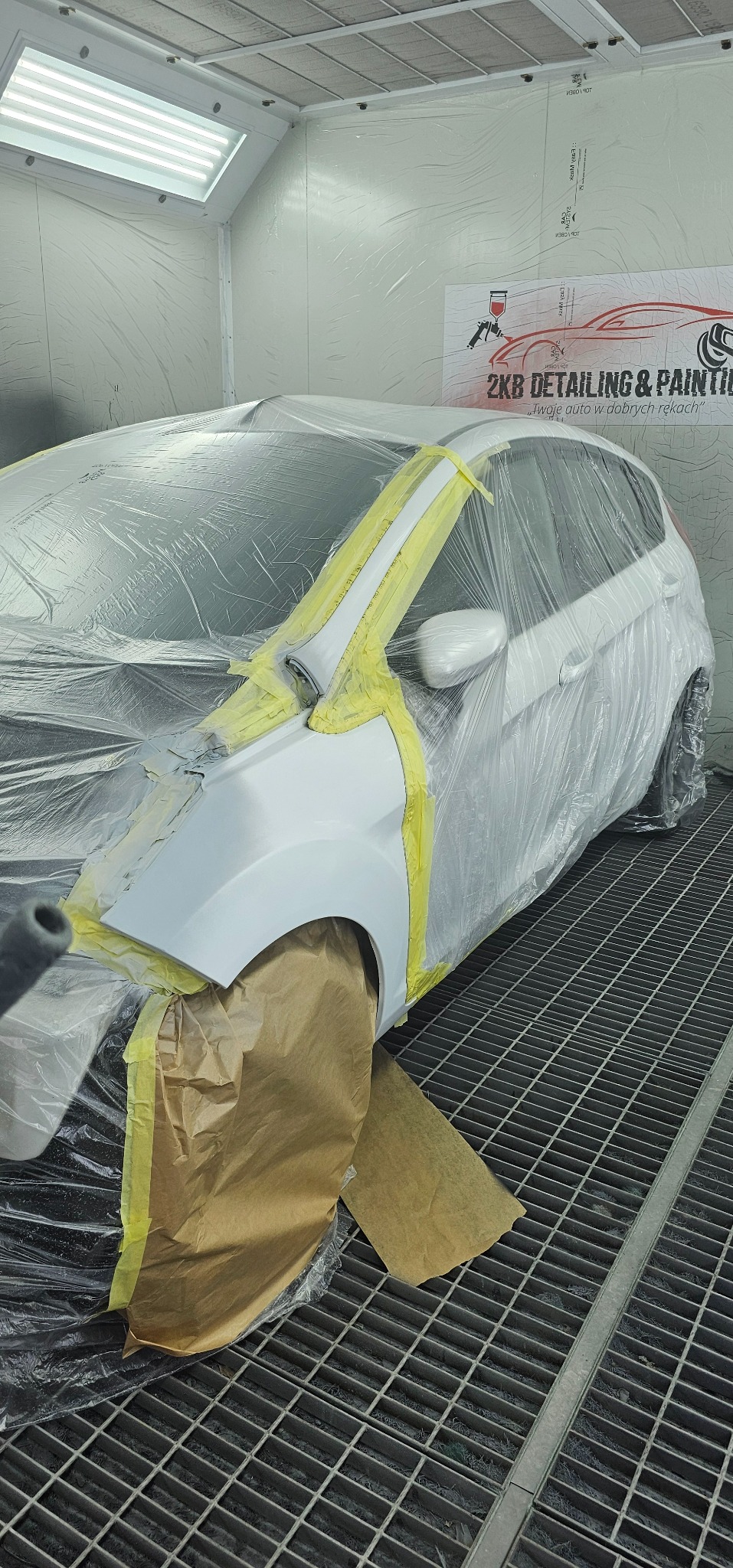 Biały samochód w komorze lakierniczej, częściowo zakryty folią ochronną i taśmą malarską, przygotowany do lakierowania; widoczna kratka podłogowa i oświetlenie sufitowe.