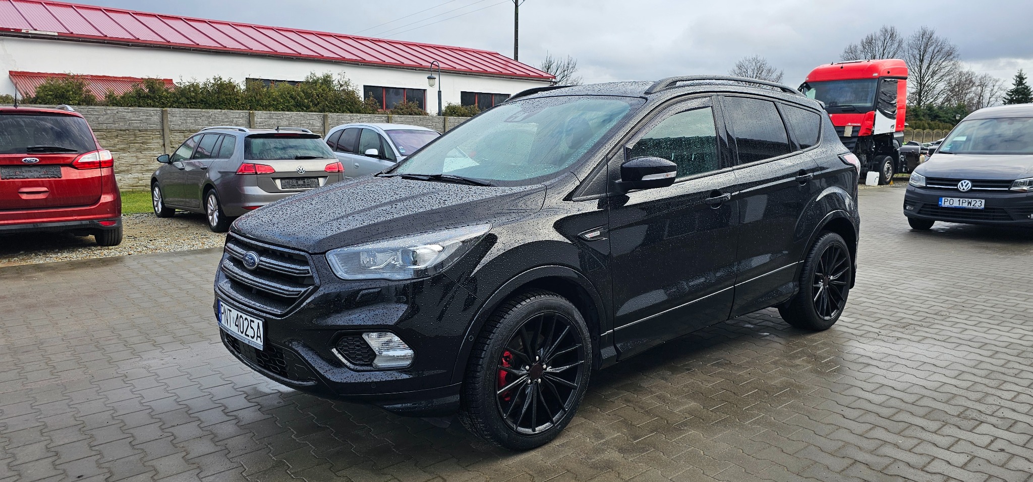 Czarny Ford Kuga po myciu, z kroplami wody na karoserii, zaparkowany na kostce brukowej, z czerwonymi zaciskami hamulcowymi widocznymi przez czarne felgi.