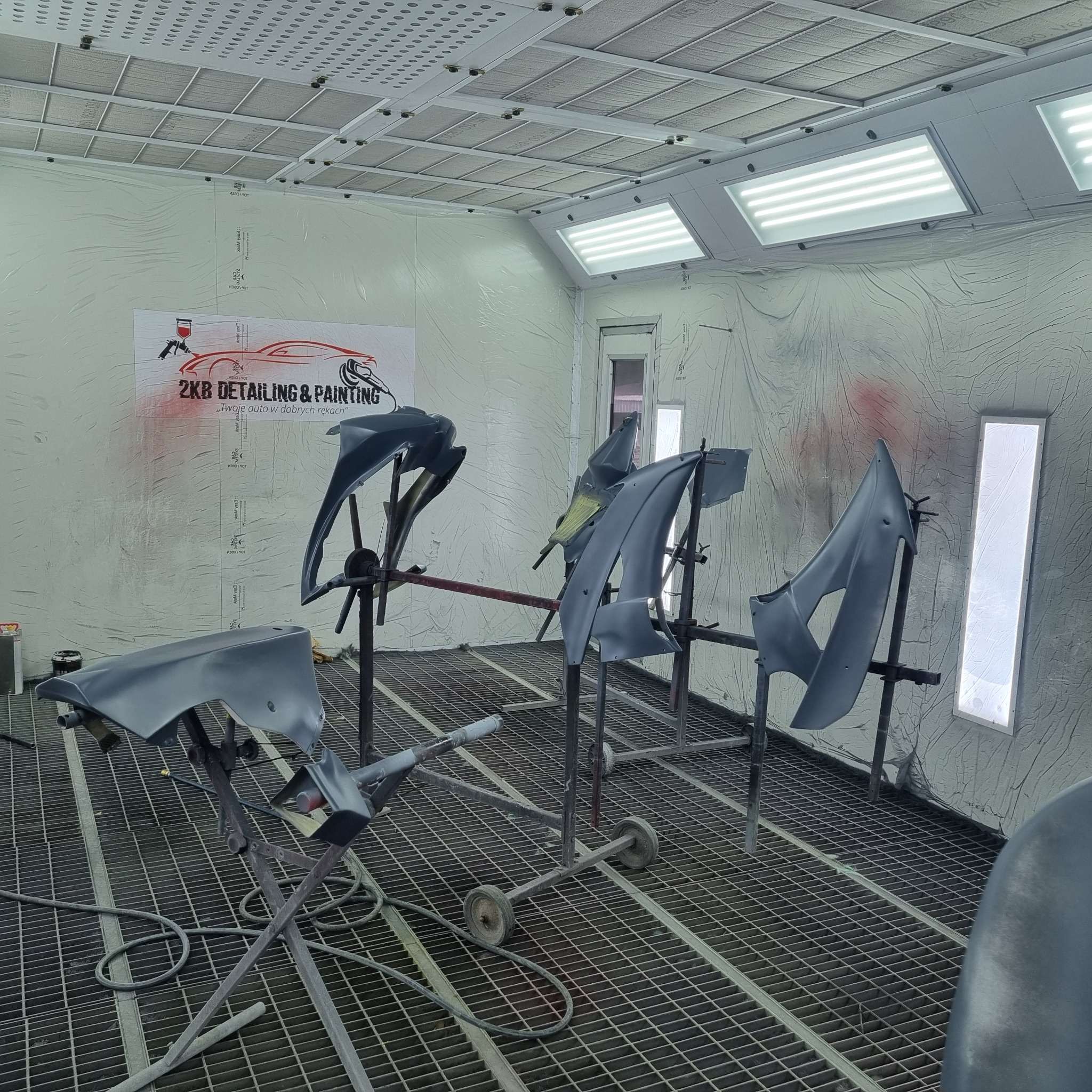 Elementy karoserii samochodowej w szarej podkładzie, zawieszone na stojakach w kabinie lakierniczej z logo firmy ZKB Detailing & Painting w tle.