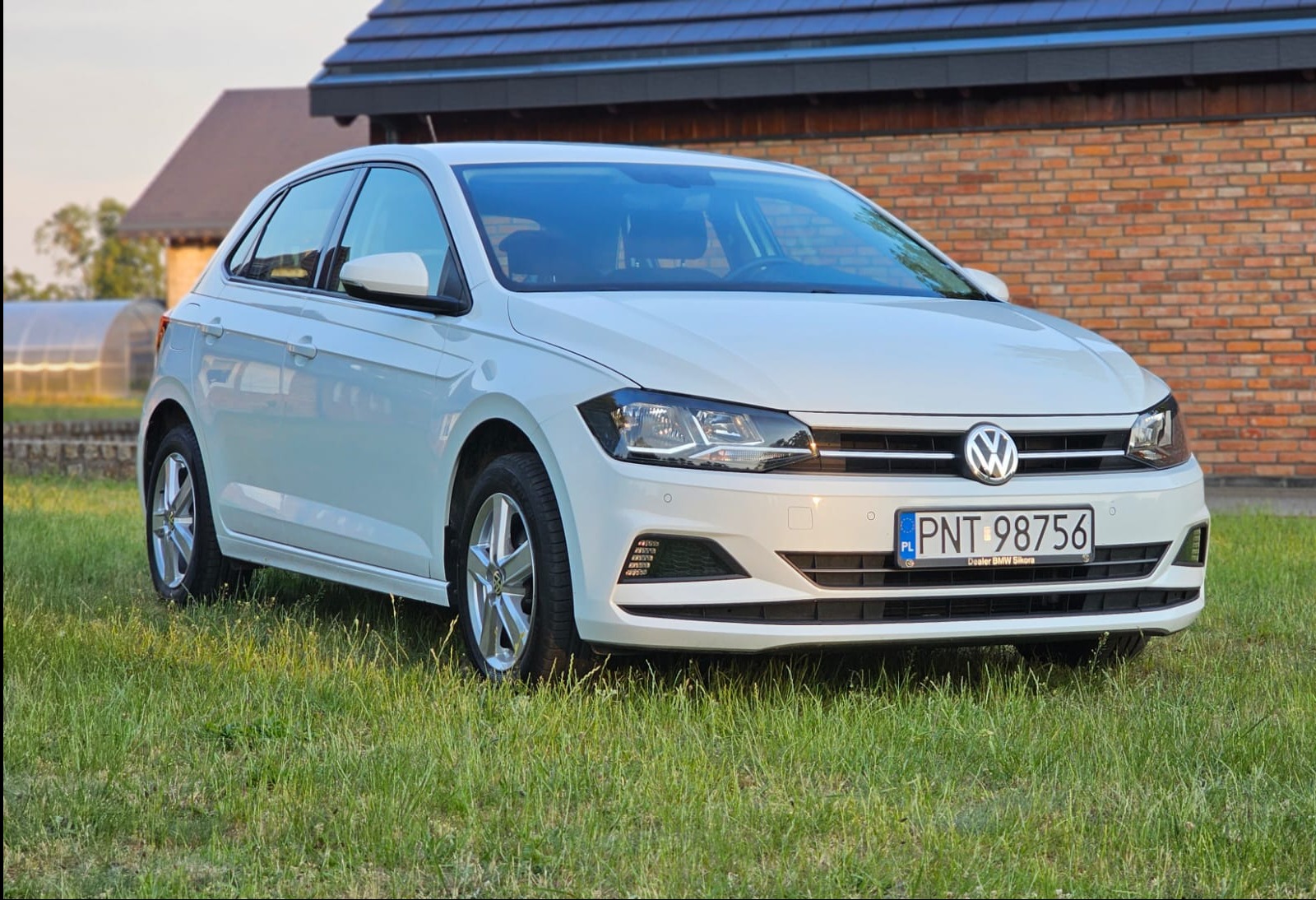 Biały Volkswagen Polo na trawie, z widocznym numerem rejestracyjnym PNT 98756 i budynkiem z cegły w tle.