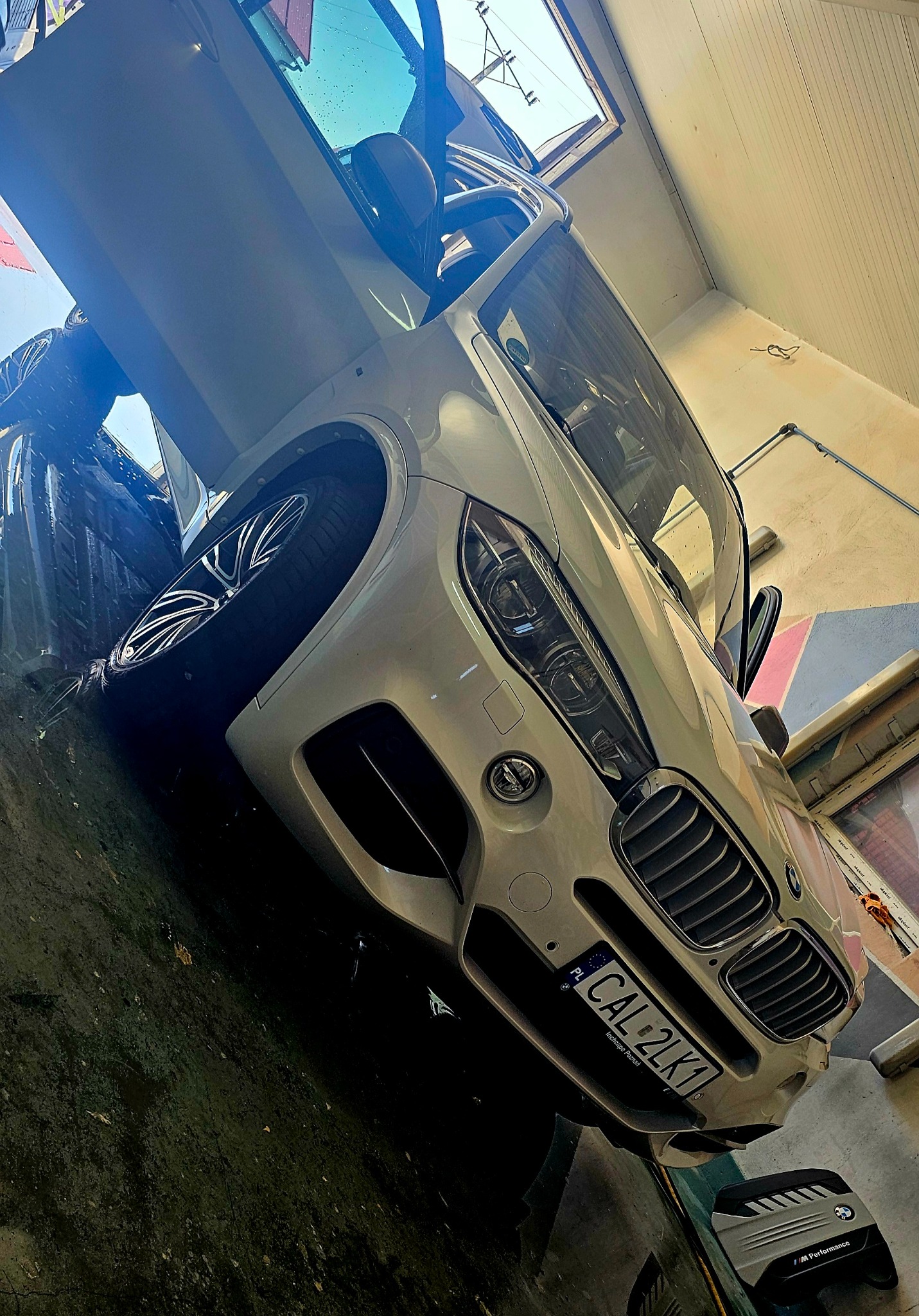 Białe BMW X5 z otwartymi drzwiami, w trakcie mycia, widoczne krople wody na karoserii, tablica rejestracyjna CAL 2LK1, wnętrze myjni z elementami instalacji.