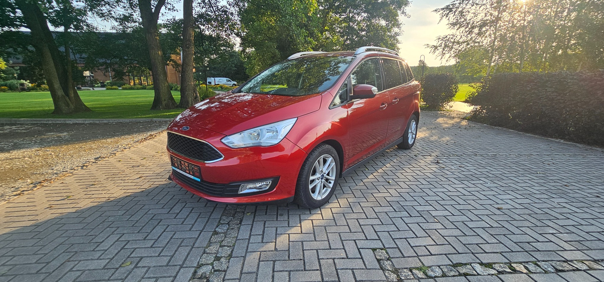 Czerwony Ford C-Max na brukowanym podjeździe w słoneczny dzień, widoczny fragment zadbanego ogrodu w tle.