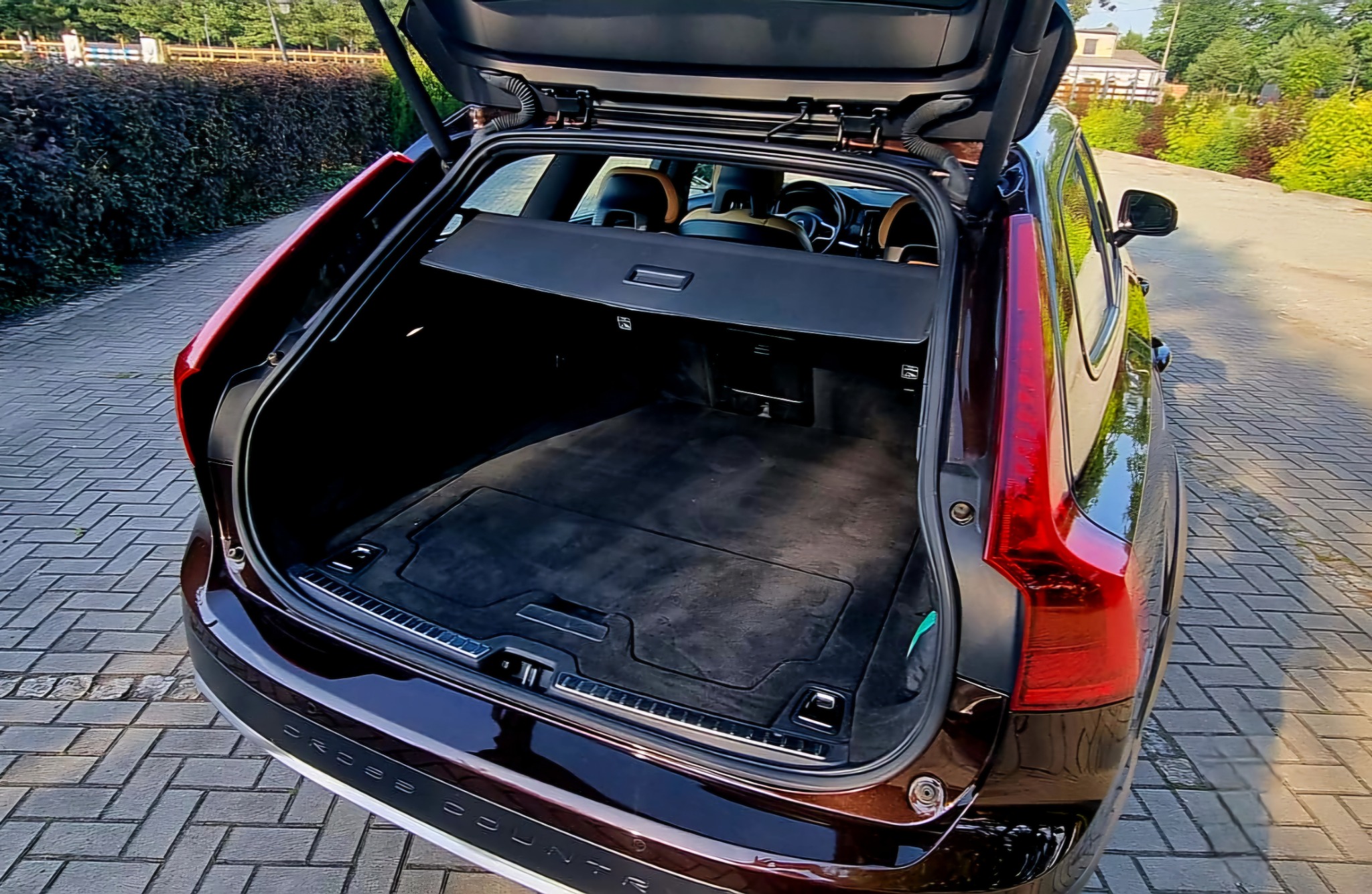 Otwarty bagażnik brązowego samochodu kombi Volvo Cross Country z widoczną czarną tapicerką i roletą bagażnika na tle brukowanej drogi i zieleni.