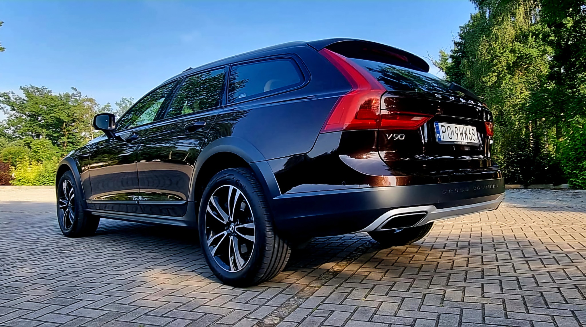 Ciemnobrązowy samochód Volvo V90 Cross Country, widziany z tyłu pod kątem, zaparkowany na kostce brukowej na tle zieleni i błękitnego nieba.