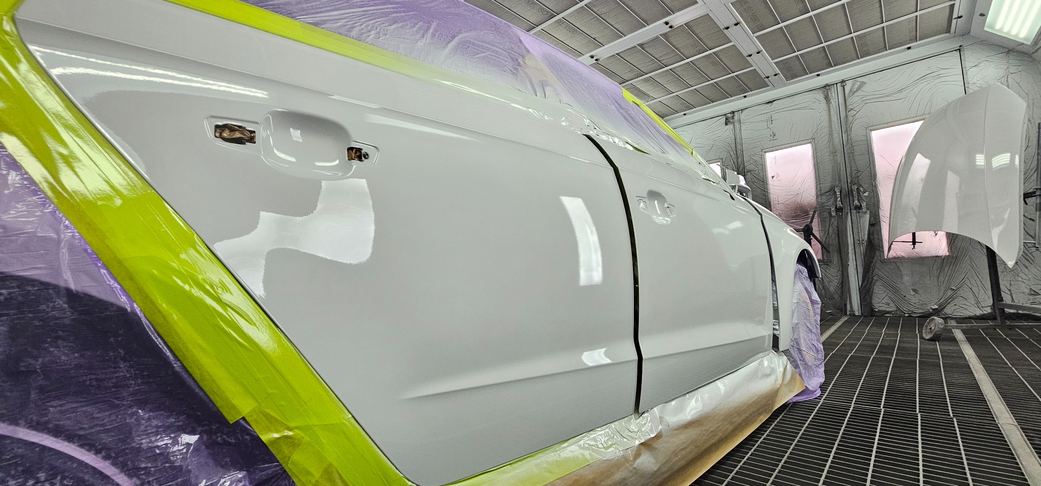 Biały samochód w kabinie lakierniczej, zabezpieczony folią i taśmą, z widoczną świeżo polakierowaną karoserią i maską czekającą na montaż.