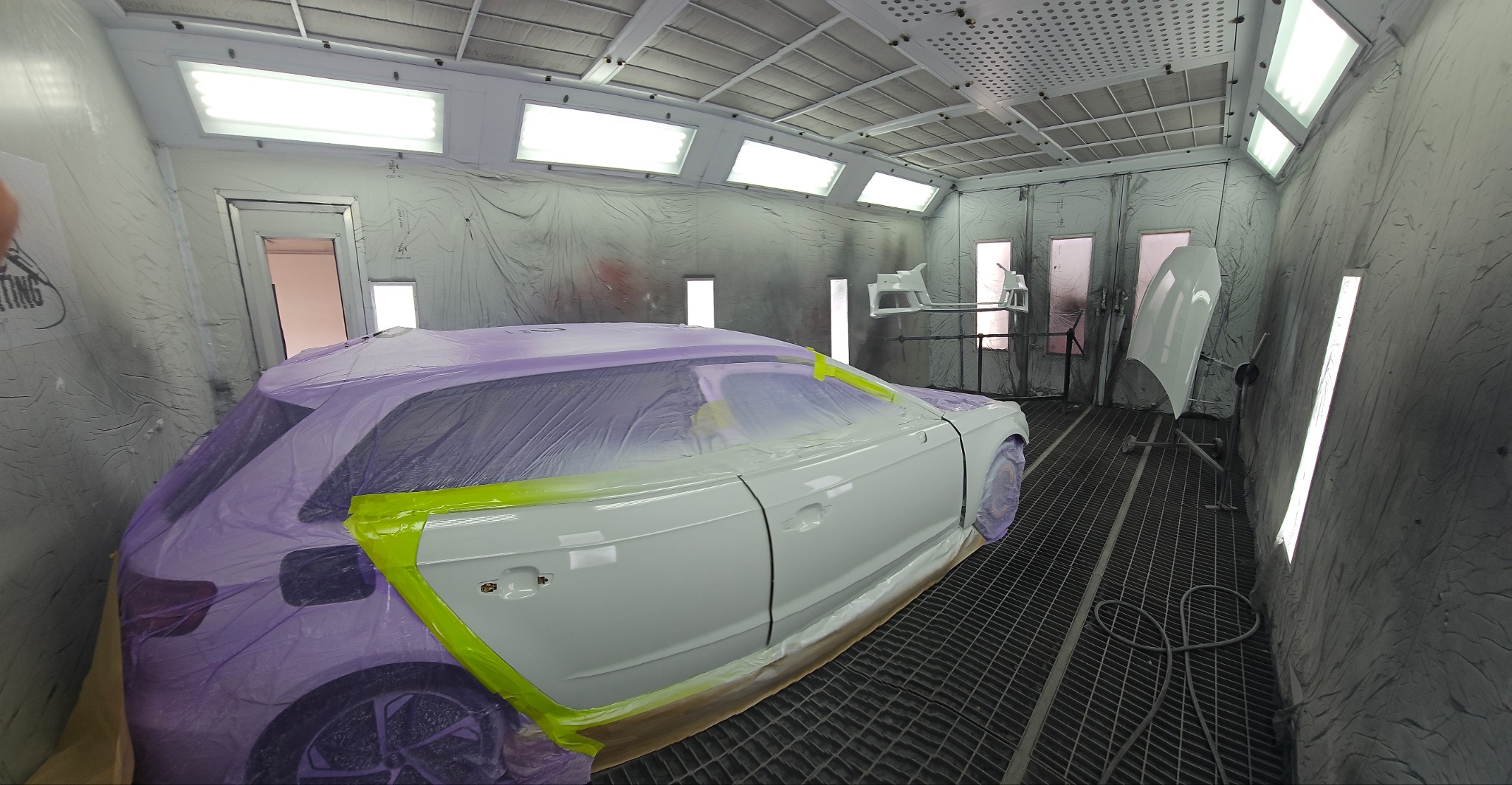 Samochód w komorze lakierniczej, częściowo oklejony folią i taśmą, przygotowany do malowania, z widocznymi elementami karoserii na stojaku w tle.