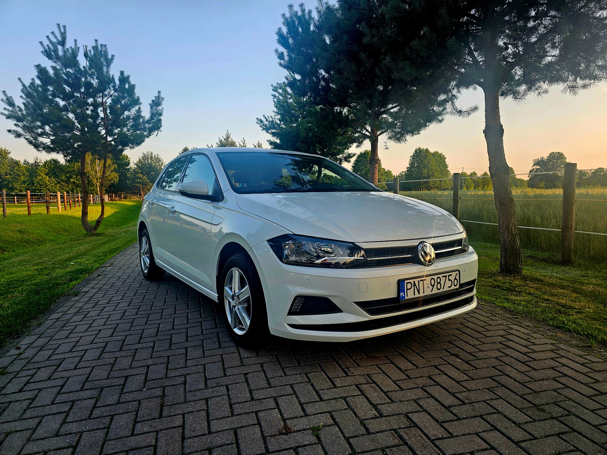 Biały samochód Volkswagen Polo zaparkowany na brukowanej drodze z widokiem na zielone drzewa i pole w tle o zachodzie słońca.