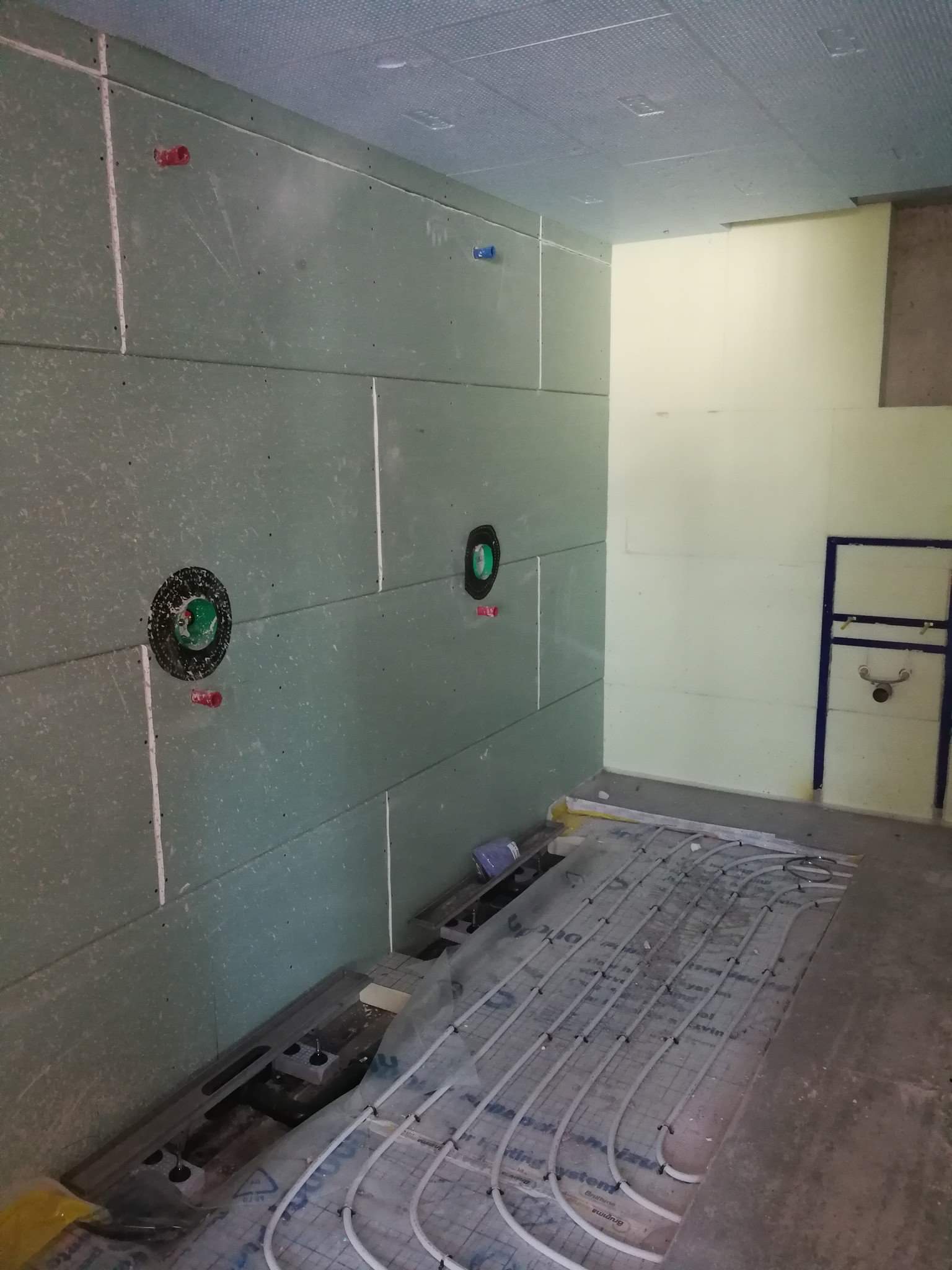 Wnętrze łazienki w trakcie montażu instalacji ogrzewania podłogowego; widoczne rury grzewcze rozłożone na folii izolacyjnej, ściany wyłożone płytami gipsowo-kartonowymi z przygotowanymi otworami...