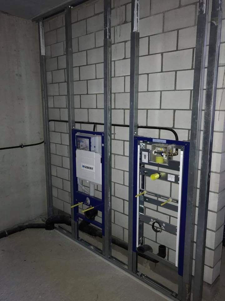 Stelaże podtynkowe Geberit zamontowane na tle surowej ściany z białej cegły, widoczne rury instalacyjne i metalowa konstrukcja nośna.