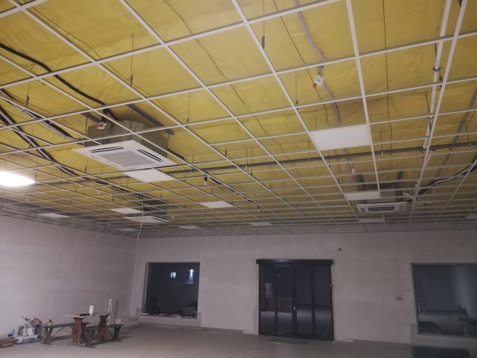 Sufit podwieszany w trakcie montażu w pomieszczeniu biurowym, widoczne instalacje, oświetlenie i jednostki klimatyzacyjne.