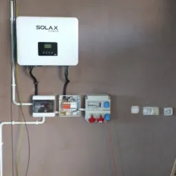 Biały falownik Solax Power zamontowany na brązowej ścianie, z widocznymi przewodami, skrzynkami elektrycznymi i gniazdkami; półka z narzędziami i materiałami budowlanymi w tle.