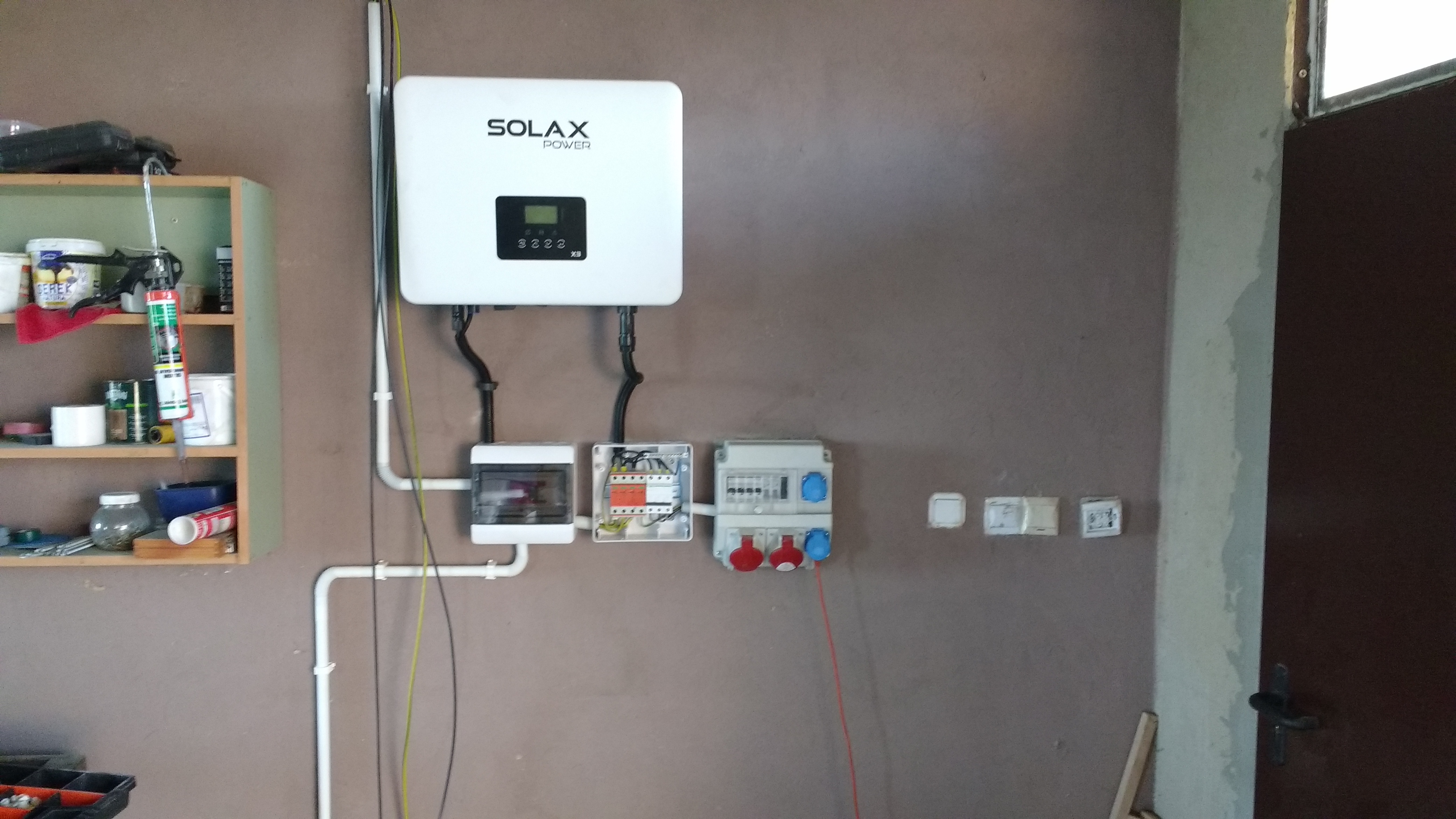 Biały falownik Solax Power zamontowany na brązowej ścianie, z widocznymi przewodami, skrzynkami elektrycznymi i gniazdkami; półka z narzędziami i materiałami budowlanymi w tle.