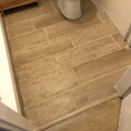 Ułożone w jodełkę, podłużne, drewnopodobne płytki podłogowe w łazience z widoczną toaletą i fragmentem szafki pod umywalkę.