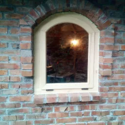 Nowe okno z jasnego drewna, łukowate, wmurowane w ścianę z czerwonej cegły z łukowym nadprożem, widoczne ślady pianki montażowej i odbicie wnętrza pomieszczenia.