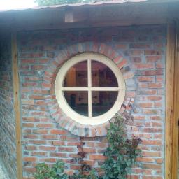 Zakład Stolarski DYL - Okrągłe okno z drewnianą ramą w murze z czerwonej cegły, z widocznymi roślinami przed budynkiem.