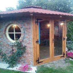 Zakład Stolarski DYL - Murowana altana ogrodowa z drewnianymi, przeszklonymi drzwiami i okrągłym oknem, otoczona roślinnością i kwiatami.