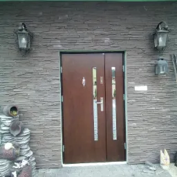 Brązowe, dwuskrzydłowe drzwi wejściowe z wąskimi przeszkleniami, otoczone elewacją z kamienia łupanego i ozdobnymi lampionami oraz dekoracjami ogrodowymi w postaci ceramicznych donic i figurek...