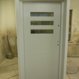 Zakład Stolarski DYL - Białe, nowoczesne drzwi z trzema poziomymi przeszkleniami, osadzone w surowym otoczeniu, widoczny fragment twarzy osoby w odbiciu szyb.