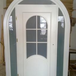 Zakład Stolarski DYL - Białe, łukowe drzwi z szybami i szprosami, widoczne w warsztacie stolarskim, z odblaskiem sylwetki osoby w szybie.