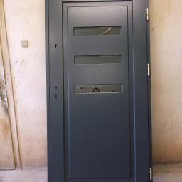 Zakład Stolarski DYL - Szare skrzydło drzwiowe z trzema przeszklonymi prostokątnymi wstawkami, widoczne zawiasy w kolorze złotym, sfotografowane na tle szarej ściany w trakcie montażu.