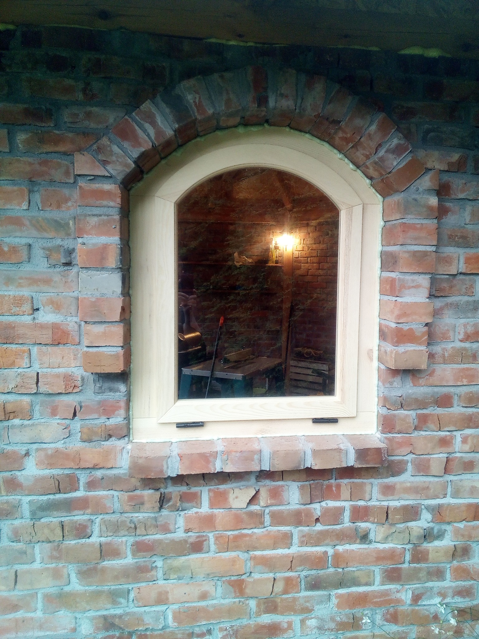 Nowe okno z jasnego drewna, łukowate, wmurowane w ścianę z czerwonej cegły z łukowym nadprożem, widoczne ślady pianki montażowej i odbicie wnętrza pomieszczenia.