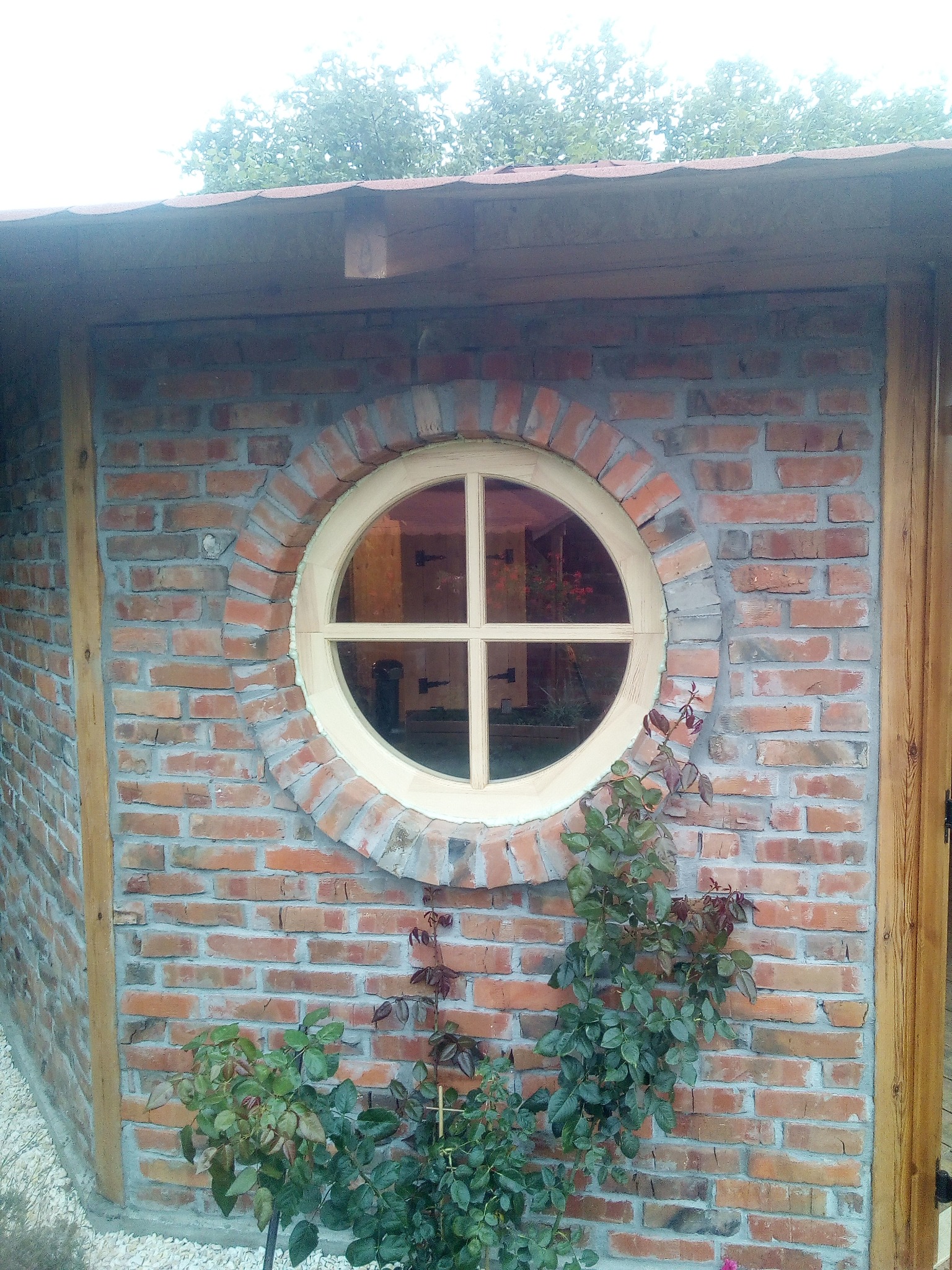 Okrągłe okno z drewnianą ramą w murze z czerwonej cegły, z widocznymi roślinami przed budynkiem.