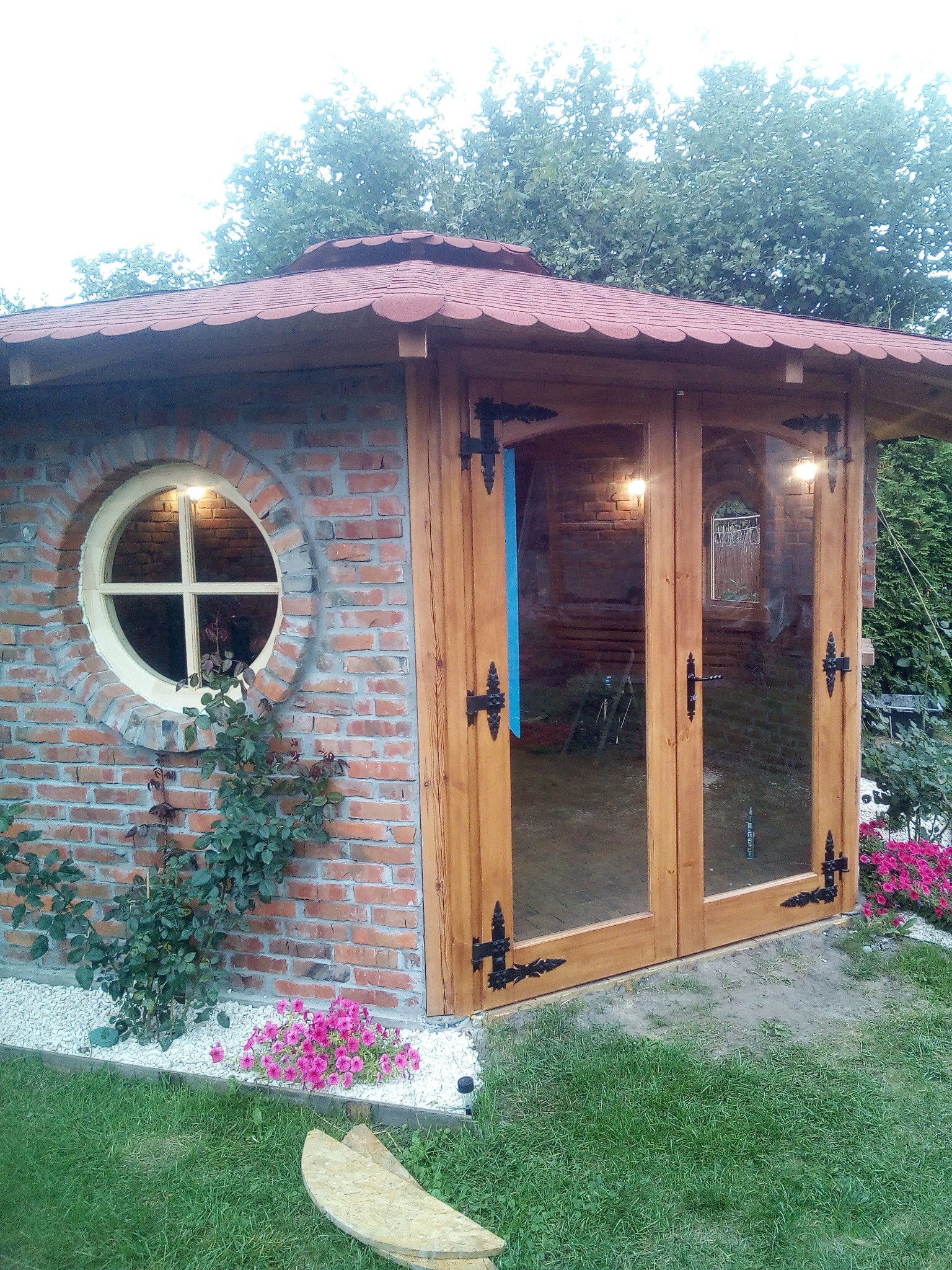 Murowana altana ogrodowa z drewnianymi, przeszklonymi drzwiami i okrągłym oknem, otoczona roślinnością i kwiatami.