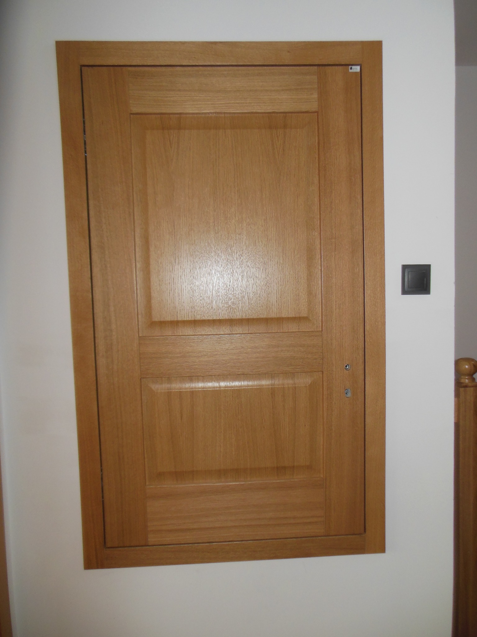 Jasna, dwupanelowa wewnętrzna konstrukcja z naturalnego drewna, zamontowana w białej ścianie, z widocznym włącznikiem światła po prawej stronie.