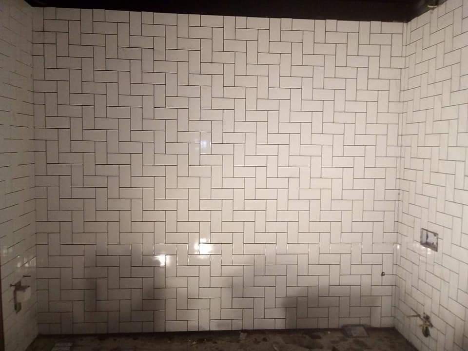 Świeżo ułożone białe, prostokątne płytki w układzie cegiełki na ścianach łazienki, z ciemnymi fugami i widocznymi elementami instalacji sanitarnych przed fugowaniem.