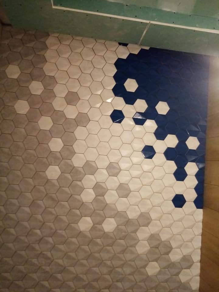 Ściana wyłożona heksagonalnymi płytkami w odcieniach szarości, bieli i granatu, ułożonymi w gradientowy wzór z przejściem kolorów, widoczne niedokończone wykończenie sufitu.