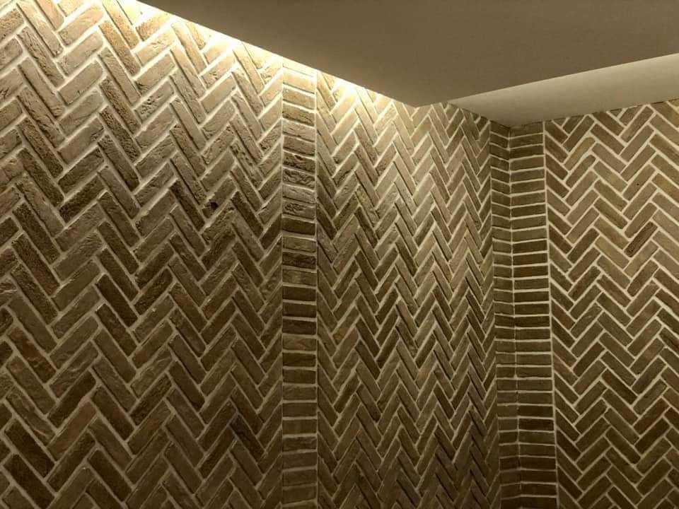 Ściana wyłożona cegłą klinkierową w jodełkę, fuga w kolorze złamanej bieli, oświetlenie górne punktowe, widok na narożnik pomieszczenia.