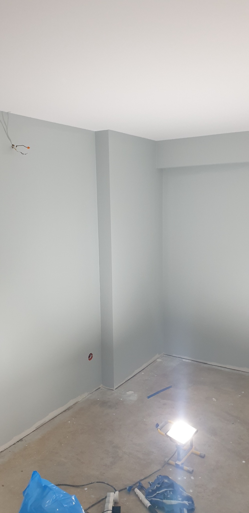 Pokój po świeżym malowaniu na szaro, z widocznymi przewodami elektrycznymi wystającymi ze ściany, lampą budowlaną na podłodze i śladami po pracach remontowych.