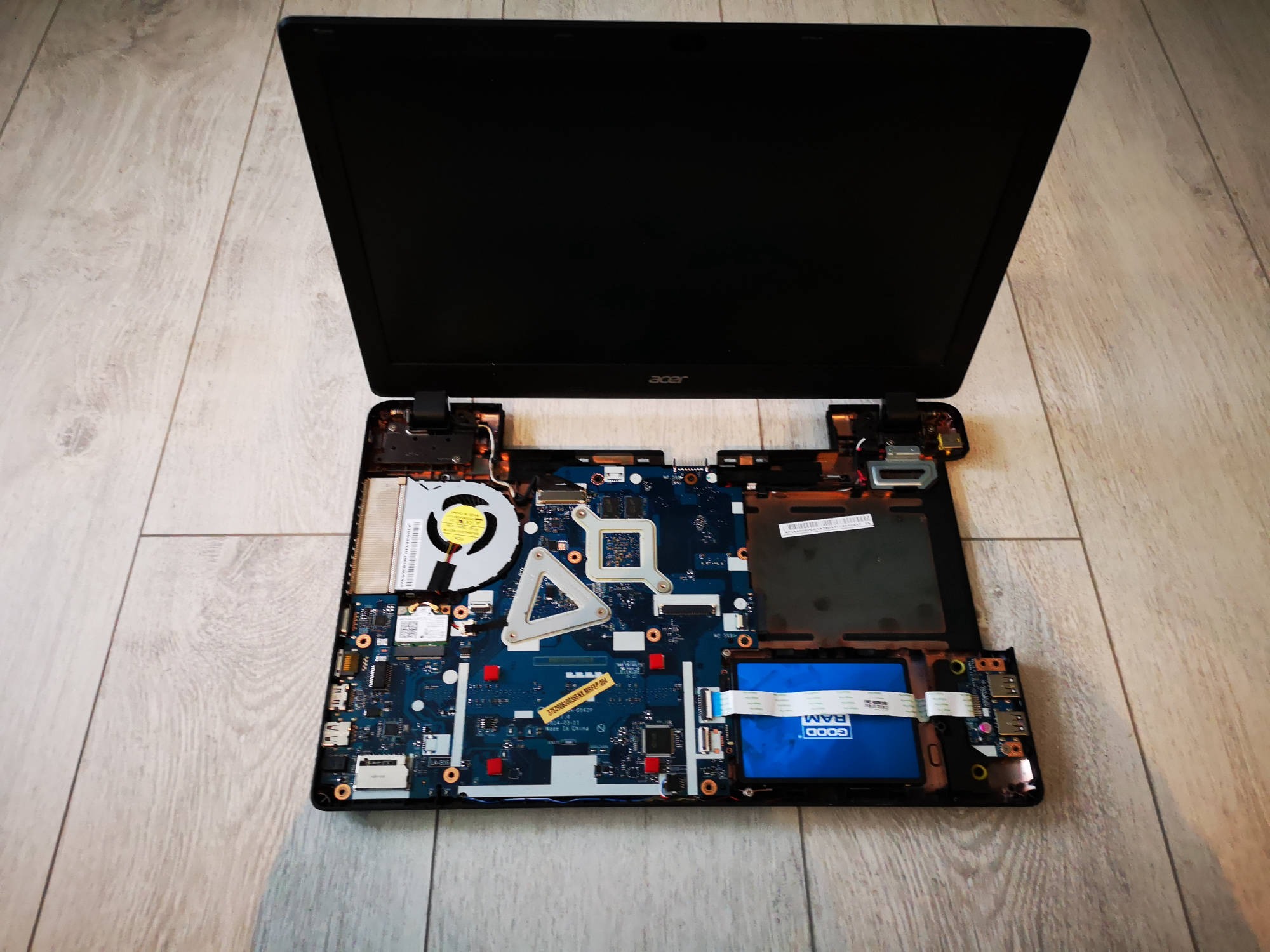Otwarty laptop Acer z widoczną płytą główną, wentylatorem i baterią, leżący na podłodze imitującej drewno.
