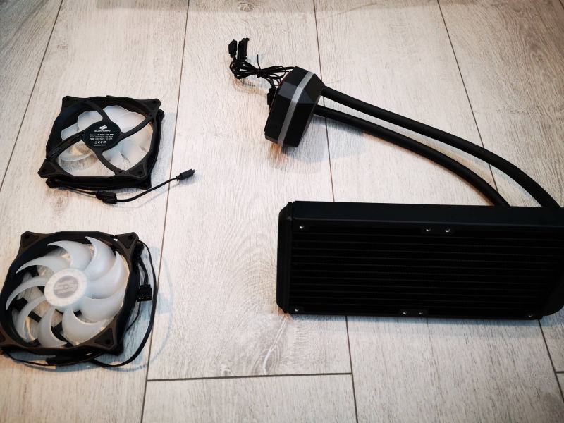 Zestaw chłodzenia wodnego do komputera, składający się z radiatora, dwóch wentylatorów i bloku wodnego z przewodami, rozłożony na podłodze imitującej drewno.