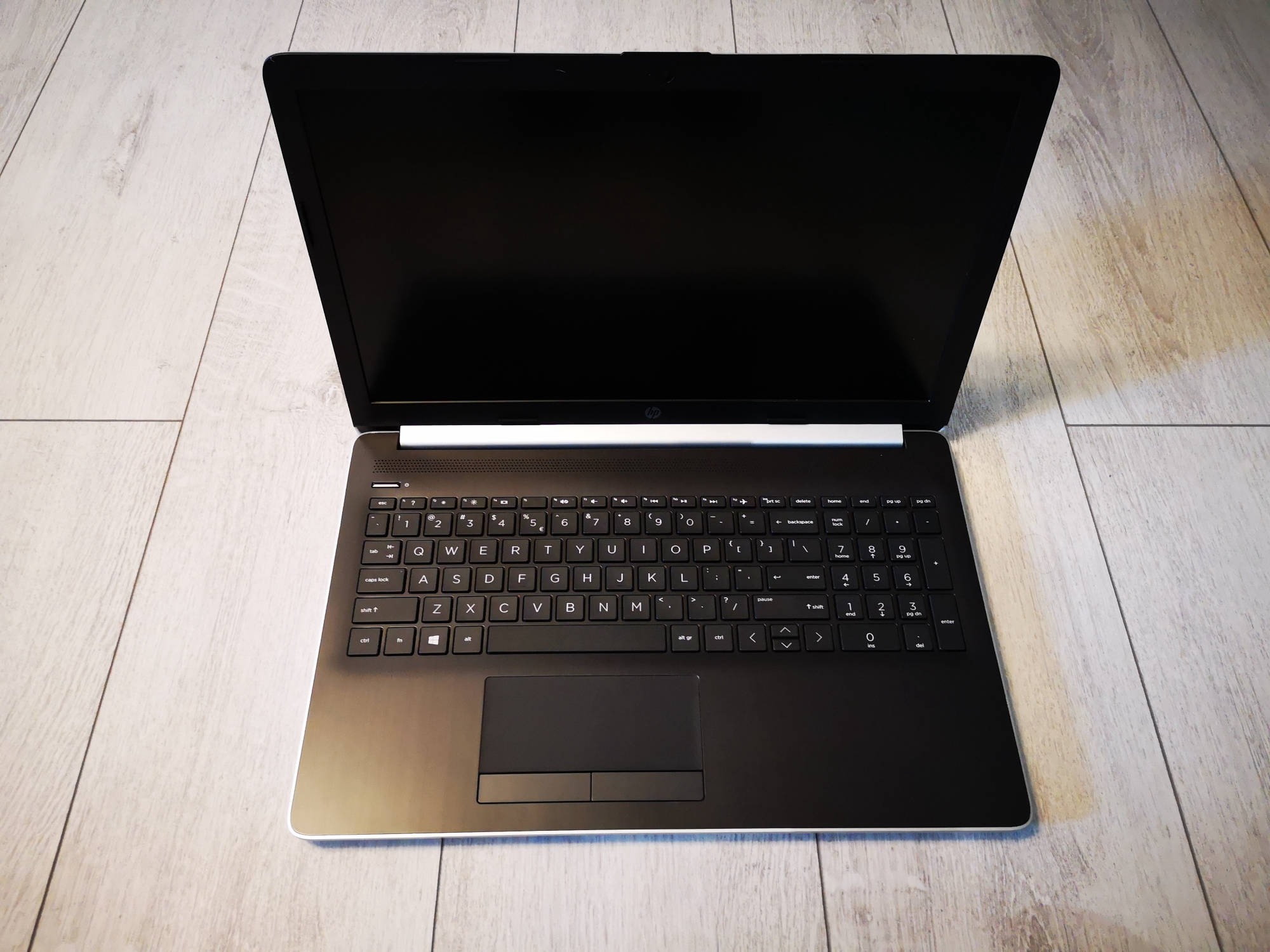 Otwarty laptop HP z czarną klawiaturą i wyłączonym ekranem, leżący na podłodze z paneli imitujących jasne drewno.