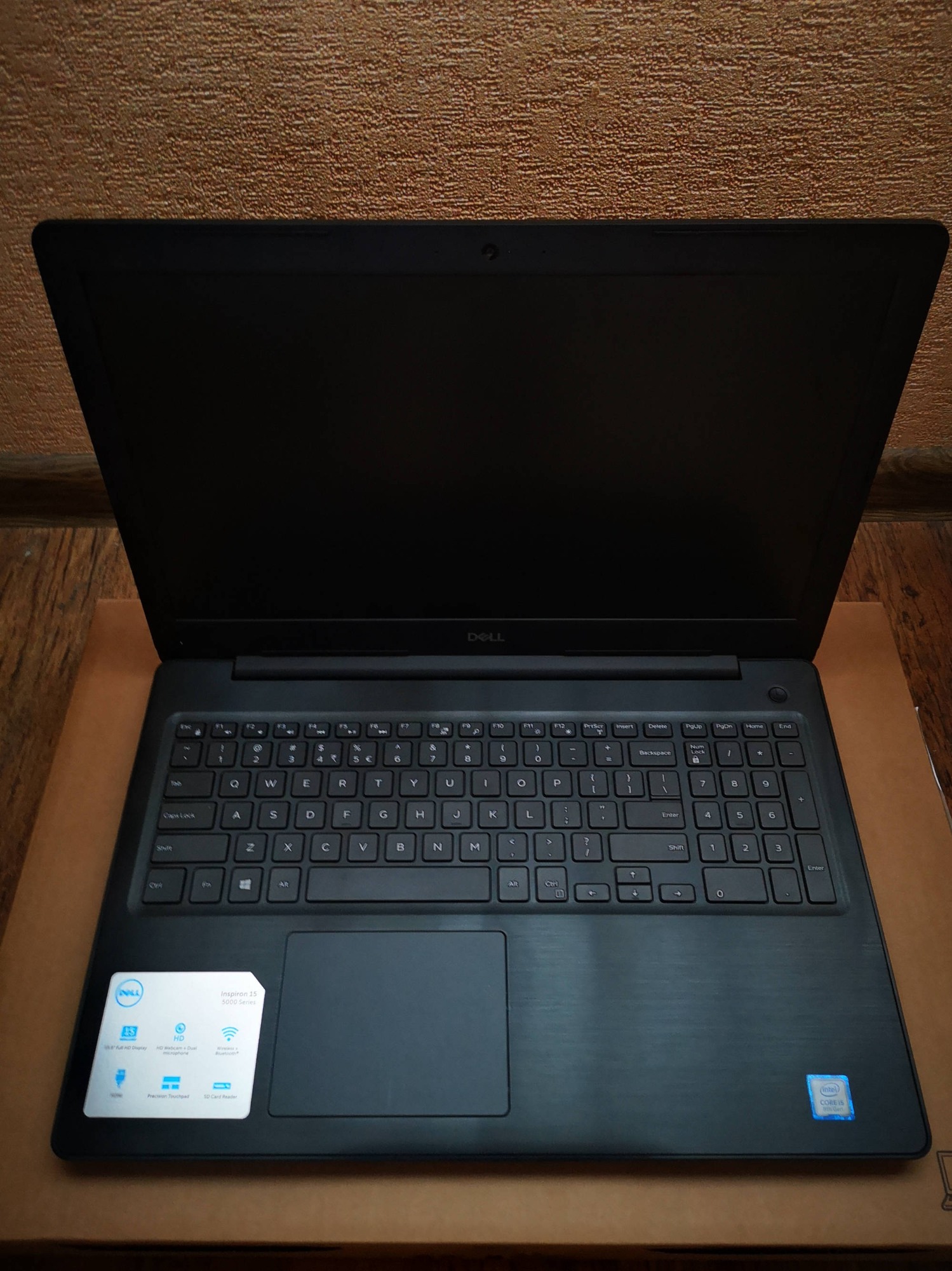 Otwarty laptop Dell Inspiron 15 5000 Series na kartonowym podłożu, widoczna klawiatura i naklejka informacyjna o parametrach.