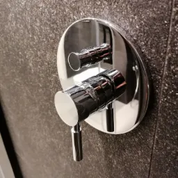 Błyszcząca, chromowana bateria prysznicowa z dwoma pokrętłami zamontowana na ciemnoszarej, teksturowanej ścianie w kabinie prysznicowej.