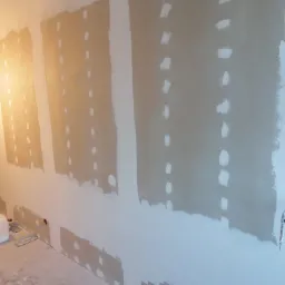 Ściana w trakcie szpachlowania płyt kartonowo-gipsowych, widoczne miejsca łączeń i wkrętów wypełnione masą, na podłodze stoją narzędzia i wiadro z zaprawą.