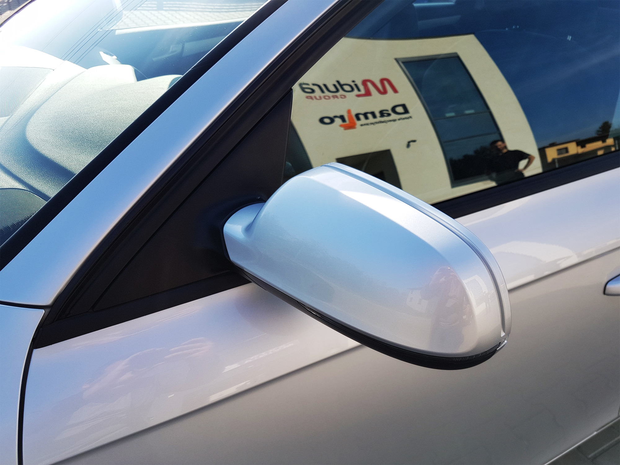 Srebrny samochód z widocznym lusterkiem bocznym, w którego odbiciu widać budynek firmy.