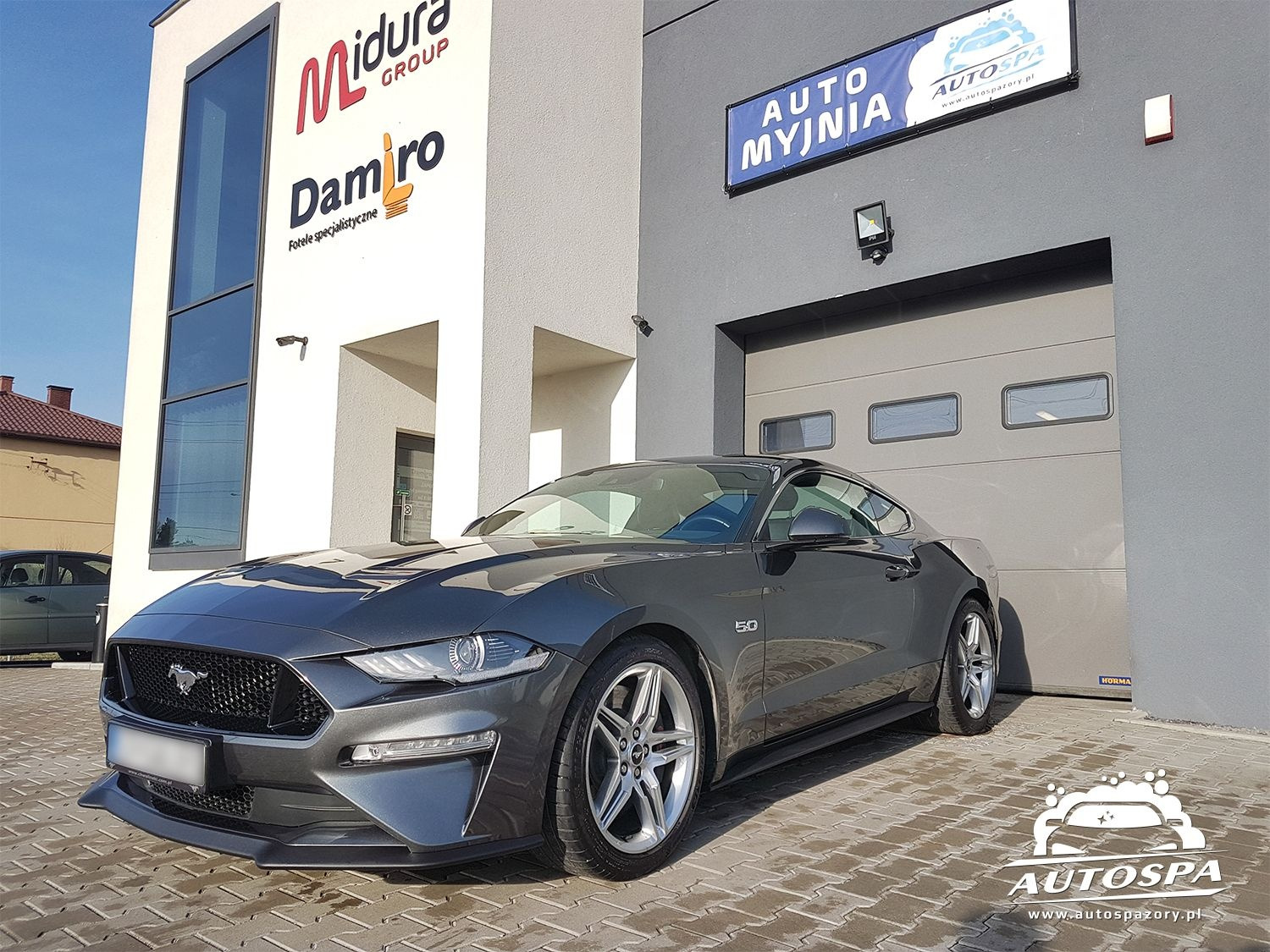 Szary Ford Mustang 5.0 zaparkowany przed budynkiem myjni samochodowej Auto Spa z widocznym logo firmy i szyldem reklamowym.